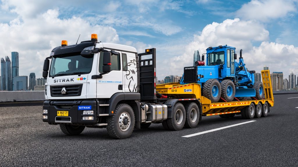 Truk Sitrak dengan trailer lowbed mengangkut motor grader